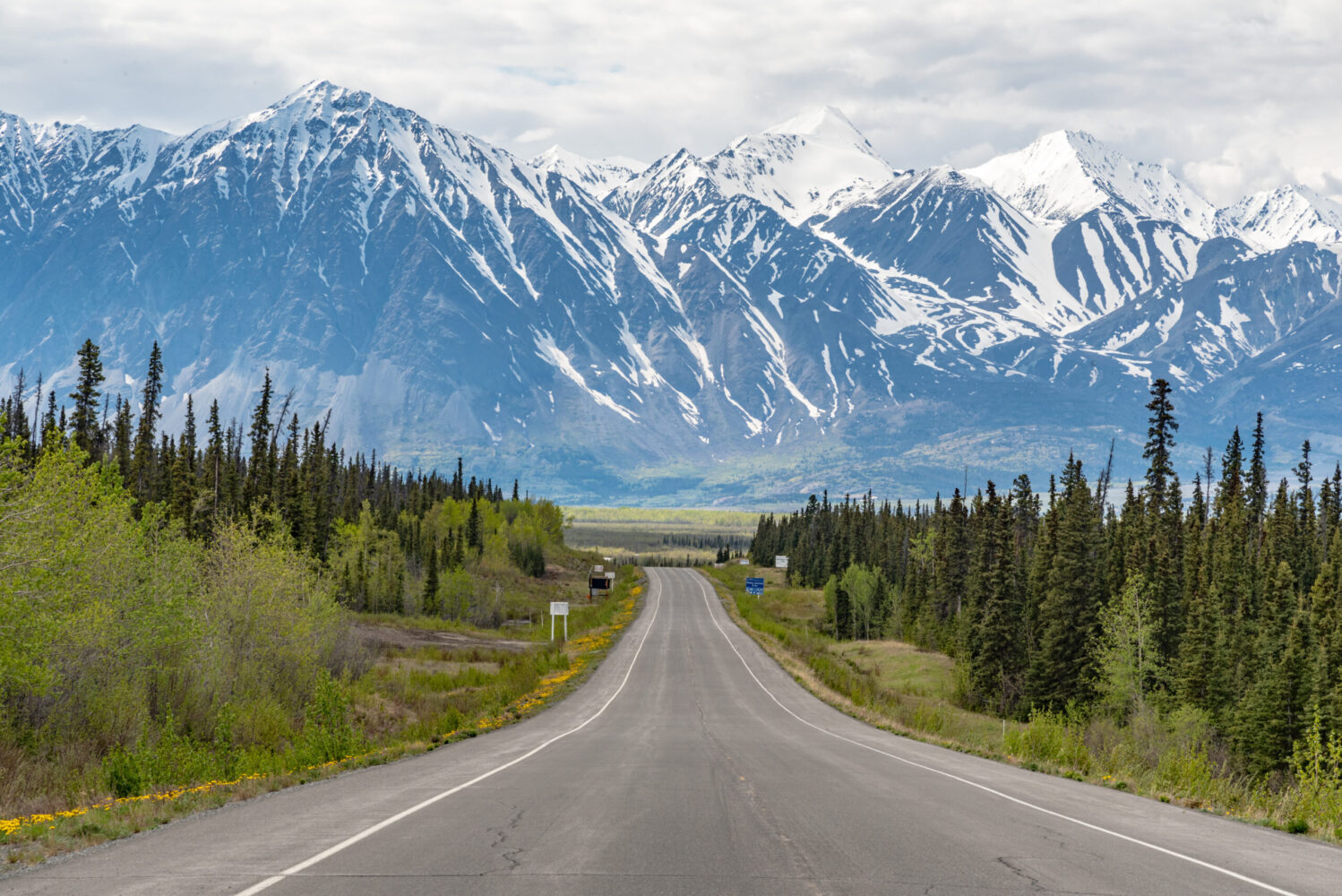 Alaska Highway driving into Haines Junction town in spring time with epic, huge mountains in far distance with amazing scenic drive ahead. Tourists, tourism shot for camping, RV, road trip.